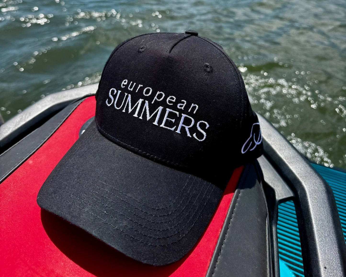 Black cap with 'European Summers' text on a boat with water and mountains in the background