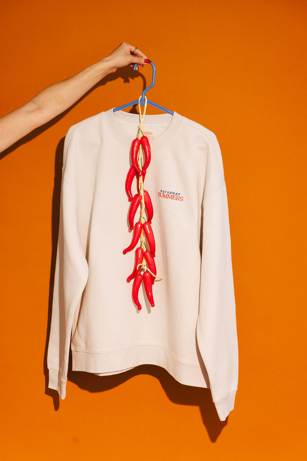 Bone colored EuroPEAN suMMERS sweatshirt with red chili pepper design on a hanger against an orange background