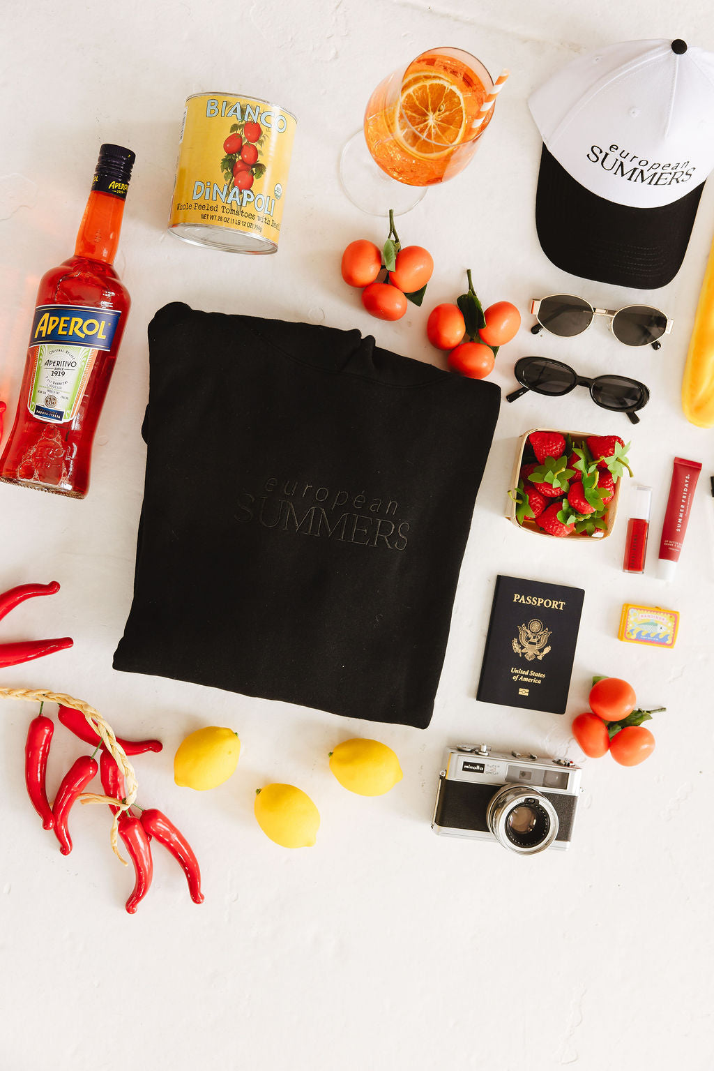 Collection of items including a bottle of Aperol, a can of Pringles, oranges, lemons, a camera, and a black bag with 'Summer's' branding on a white surface.