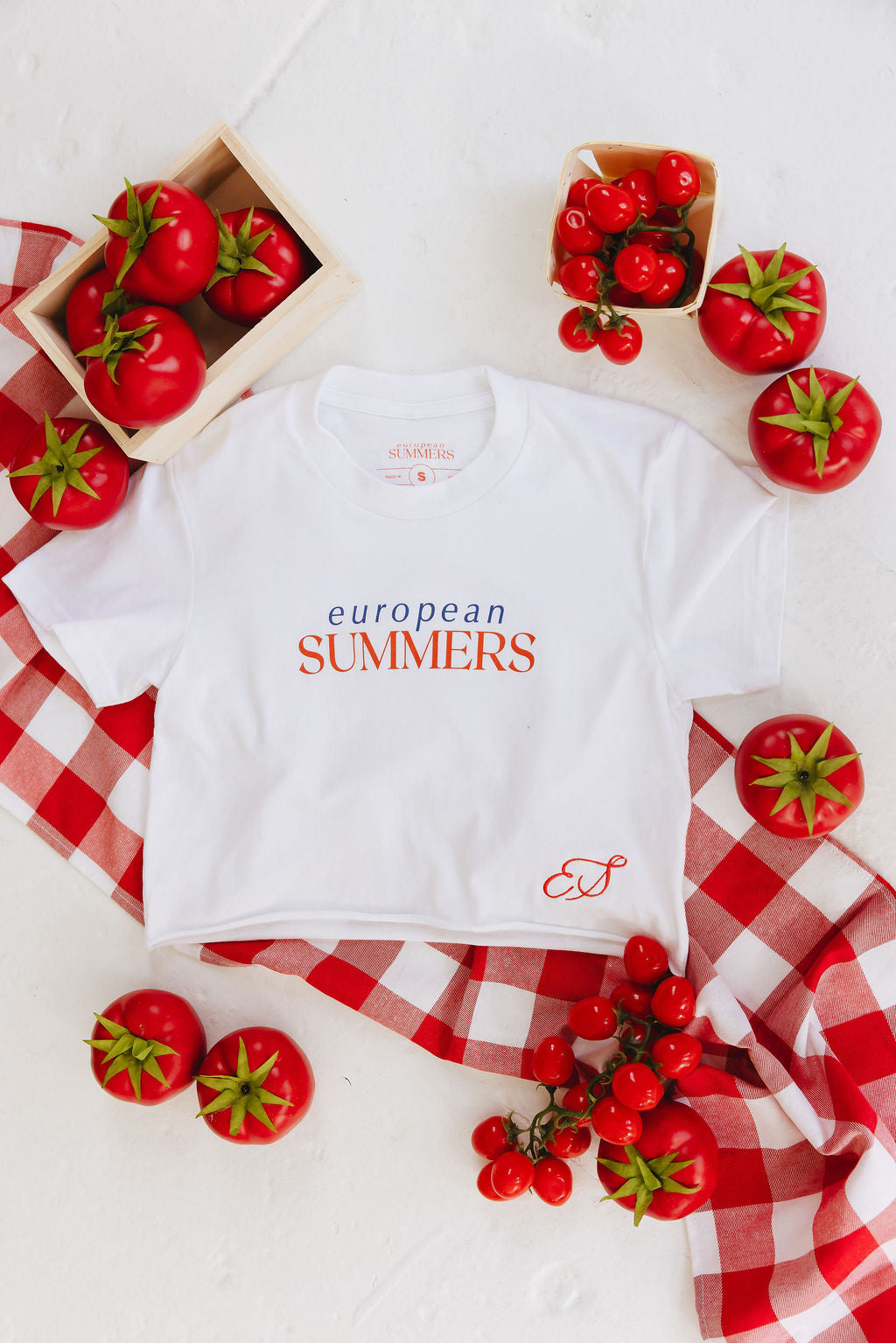 White baby t-shirt with 'European Summers' text on a white background with red tomatoes and checkered fabric.
