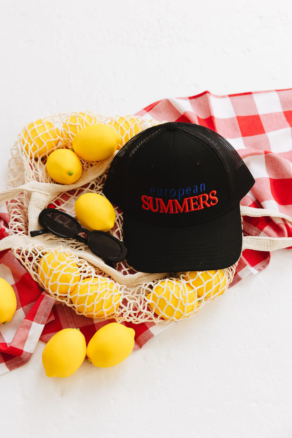 Black cap with 'European Summers' text on a white surface with lemons and a checkered cloth.