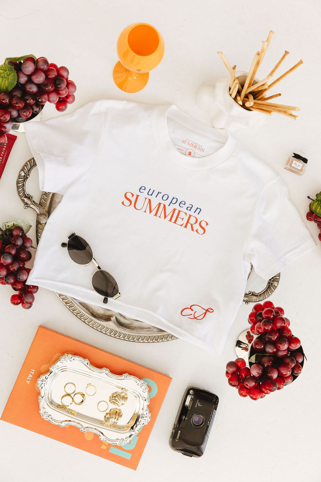 White t-shirt with 'European Summers' text on a table with grapes, books, and a camera.