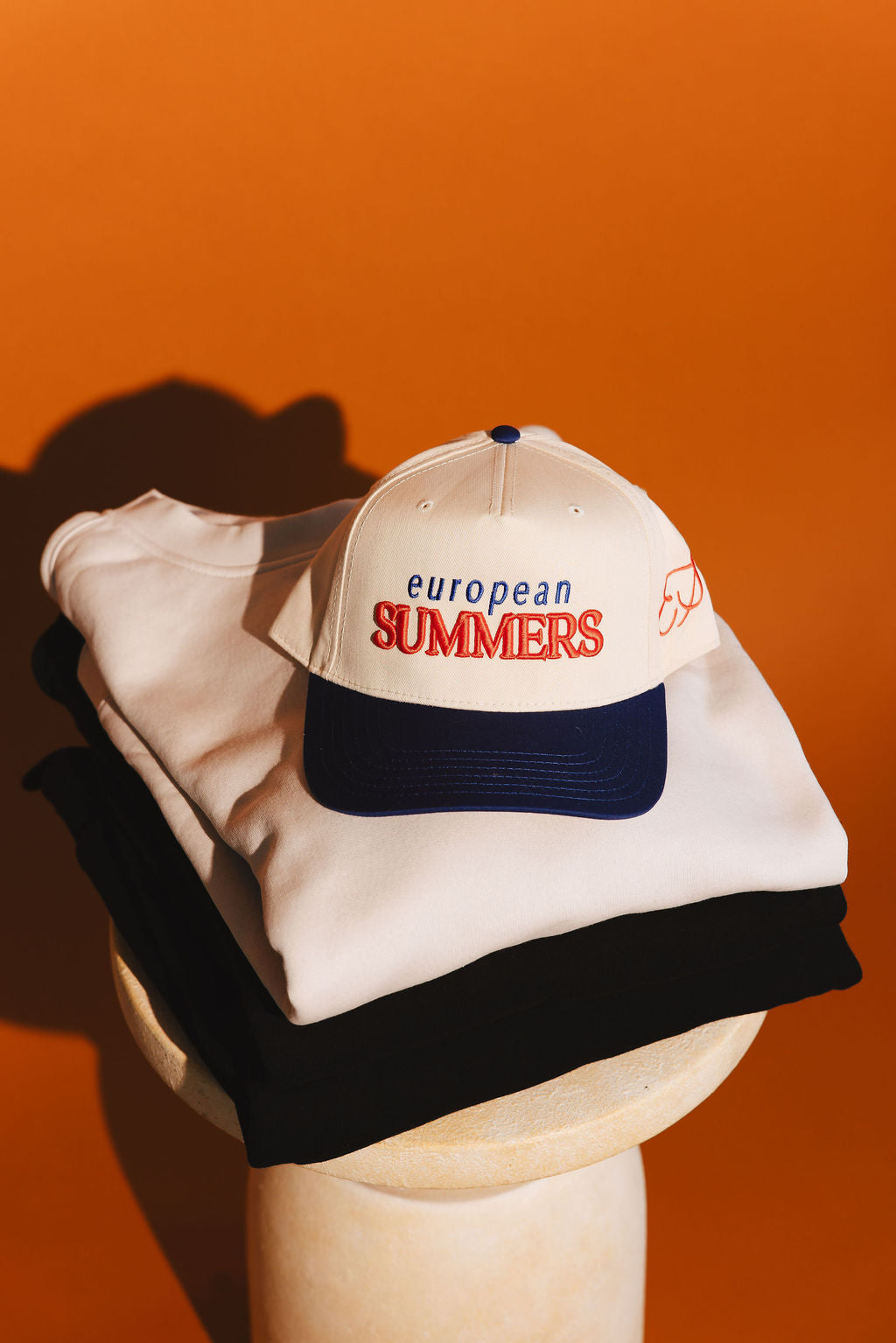 White cap with 'European Summers' text on a white surface against an orange background and crewneck layers