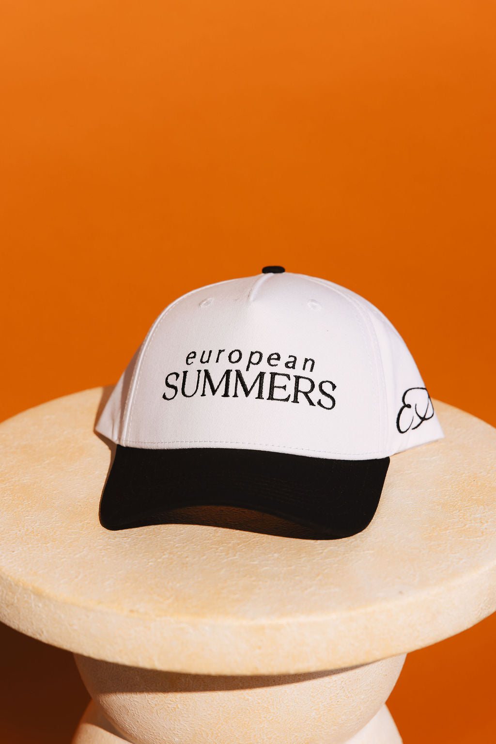 White cap with 'European Summers' text on a lifestyle wooden stand against an orange background