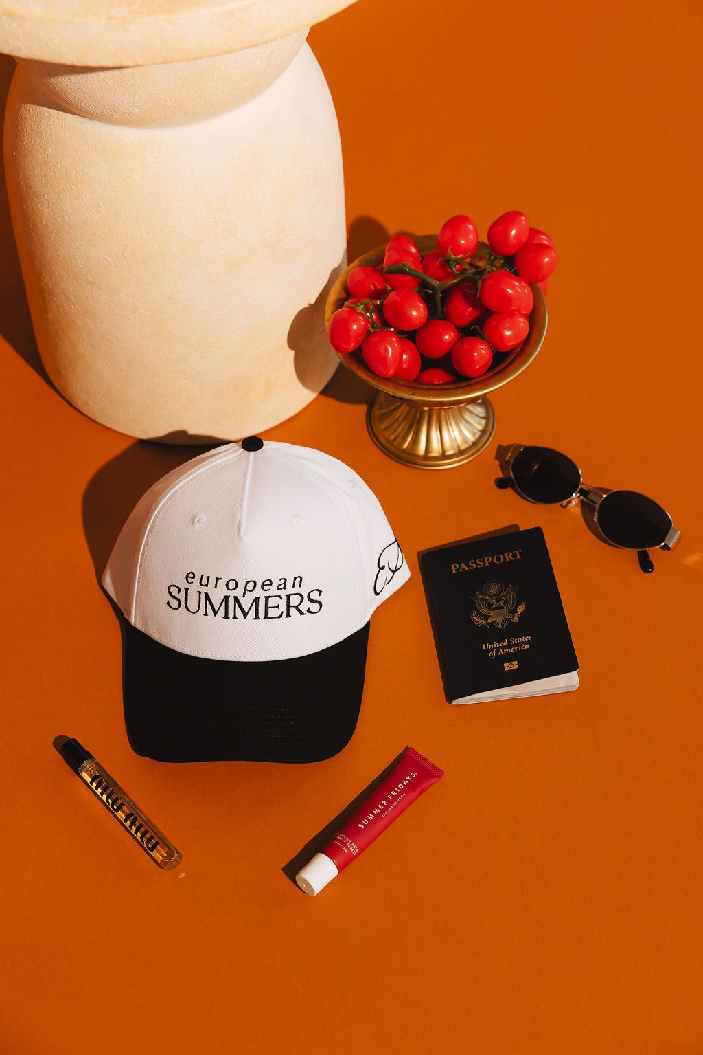 Cap with 'European Summers' text, passport, sunglasses, and lip products on an orange surface.